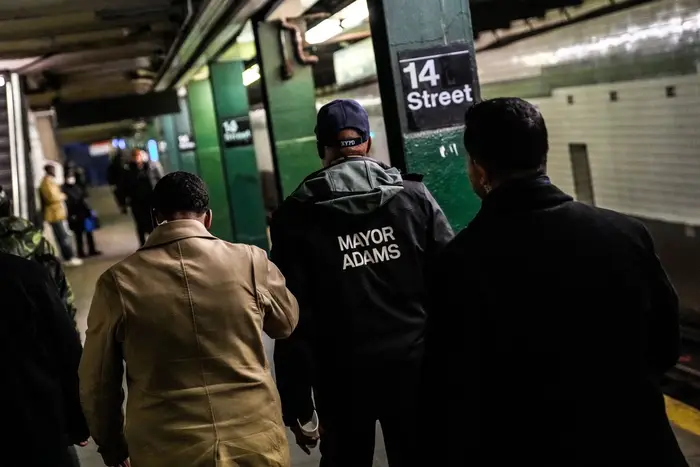 Mayor Eric Adams, flanked by two aides, walks down a subway platform. He is wearing a jacket with the words "Mayor Adams" printed on the back.